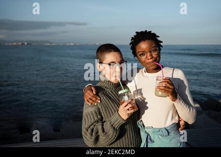 Junge Frauen trinken Fruchtsaft, während sie gegen das Meer stehen Stockfoto