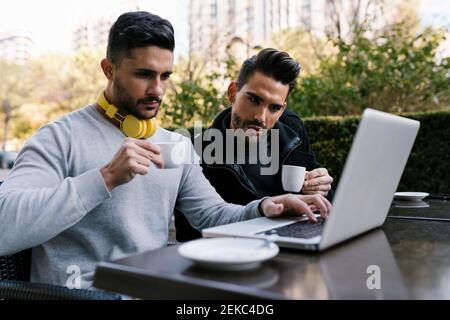 Männliche Freunde schauen auf Laptop, während mit Kaffee sitzen an Straßencafé an der Straße Stockfoto