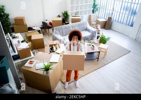 Glückliche junge Afro-Frau, die Karton trägt, während sie neu in Bewegung ist Zu Hause Stockfoto