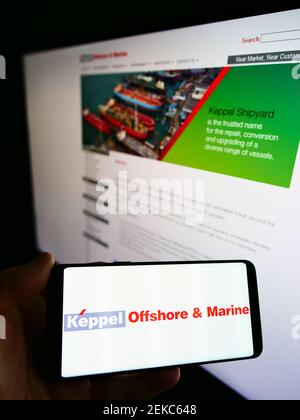 Person hält Handy mit Logo der singapurischen Firma Keppel Offshore und Marine auf dem Bildschirm vor der Webseite. Fokus auf Handy-Display. Stockfoto