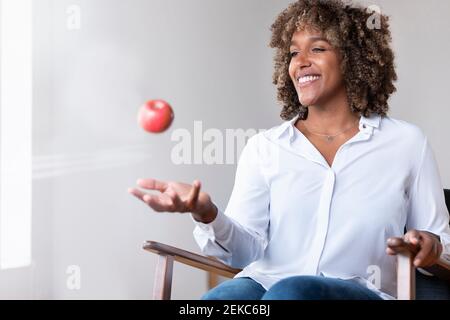 Frau lächelt, während sie mit Apfel auf dem Sessel sitzt Zu Hause Stockfoto