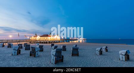 Deutschland, Mecklenburg-Vorpommern, Heringsdorf, Kapuzenliegen am leeren Strand in der Abenddämmerung mit beleuchtetem Ahlbeck Pier im Hintergrund Stockfoto