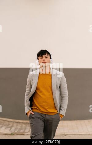Modischer Mann mit Kopfhörern, der mit den Händen in den Taschen gegen den Fuß geht Wand Stockfoto