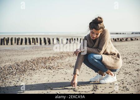 Schöne Frau zieht Herz auf Sand gegen Himmel am Strand Stockfoto