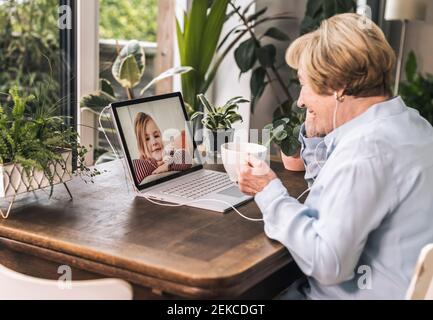 Lächelnde Großmutter auf Videoanruf mit Enkelkind durch Laptop während Mit schwarzem Kaffee zu Hause Stockfoto