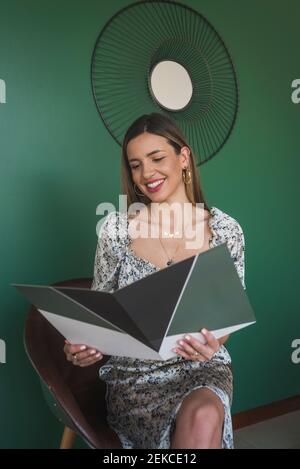 Lächelnde Geschäftsfrau lesen Datei, während auf Stuhl gegen grün sitzen Wand Stockfoto