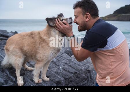Glücklicher Mann, der einen Hund umarmt, der auf einer Felsformation am Strand steht Stockfoto