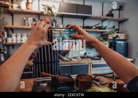 Junge Frau filmt reifen Koch durch Handy, während stehend In der Küche Stockfoto