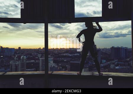 Silhouette eines Mädchens, das am Rande des Gebäudes gegen den Sonnenuntergang im städtischen Stadthintergrund steht. Top-Level-Konzept Stockfoto