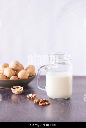 Pflanzliche Walnussmilch in einer Glasschale mit einem Griff. Walnüsse in einer Platte im Hintergrund. Gesunde Getränke, Vegetarismus. Seitenansicht, vertikales Foto. Stockfoto