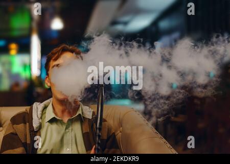 Junger Mann im Café raucht Shisha. Dicker Rauch von Shisha. Mann auf dem Sofa im Restaurant. Neonschilder auf dem Hintergrund. Entspannen Sie sich in der Shisha Bar. Stockfoto