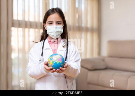 Kleines Kind, gekleidet als Arzt, mit einer medizinischen Maske, die einen Ball der Welt in den Händen hält. Konzept des Weltgesundheitstages. Leerzeichen für Text. Stockfoto