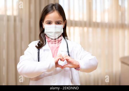 Porträt eines kleinen Kindes als Arzt gekleidet und eine medizinische Maske bilden ein Herz mit ihren Händen. Konzept von Gesundheit und Wellness. Leerzeichen für Text. Stockfoto