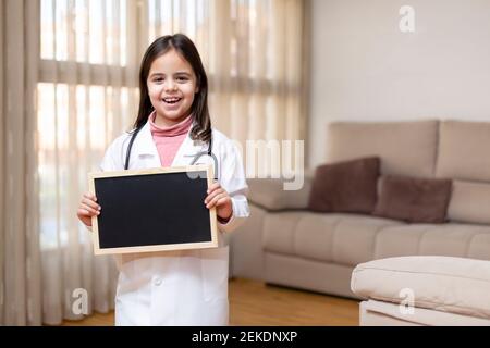 Lächelndes kleines Kind in Arztuniform, das zu Hause eine Tafel hält. Leerzeichen für Text. Stockfoto