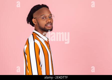 Half Turn Portrait stilvoller afrikanischer Mann mit Dreadlocks und Ohrpiercing in gestreiftem Hemd, der mit ruhiger, selbstbewusster Ausdruckskraft auf die Kamera blickt. Stehbolzen für den Innenbereich Stockfoto