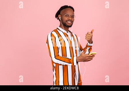 Glücklich zufrieden afro-amerikanischen Mann in Kopfhörer mit Dreadlocks im gestreiften Hemd zeigt Daumen nach oben mit mobilen Anwendung, um Musik zu hören. Innen Stockfoto