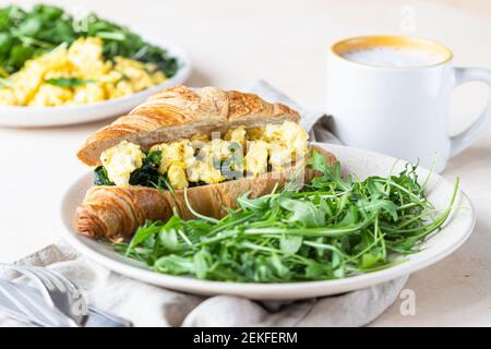 Köstliches Croissant-Sandwich mit Rührspinat-Eiern serviert mit Rucola auf dem Teller und Kaffeetasse, heller Betonhintergrund. Selektiver Fokus. Stockfoto