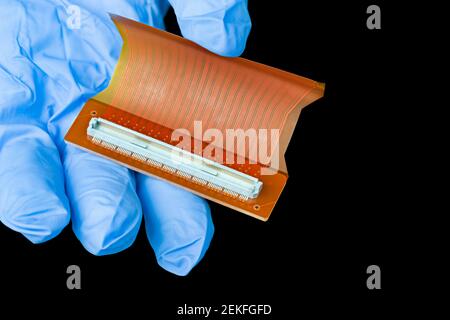 Elektronische flexible Leiterplatte in der Hand des Technikers auf schwarzem Hintergrund isoliert. Finger Detail in blauen Handschuh hält orange Kunststoff PCB. Weißer Anschluss. Stockfoto