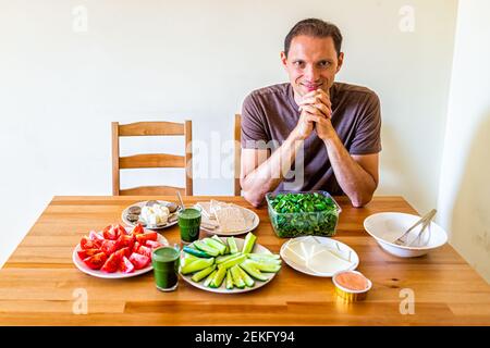 Glücklicher junger Mann sitzt auf Stuhl von Holztisch mit Einstellung von gesunden veganen vegetarischen Mittag-oder Abendessen grünes Gemüse Saft zu Hause Stockfoto