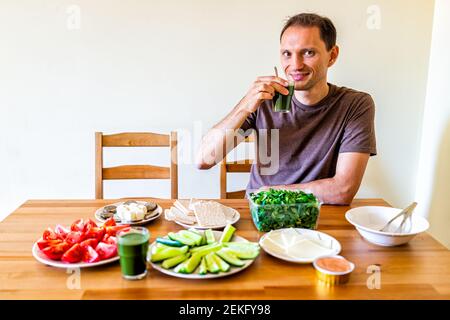 Glücklicher junger Mann sitzt auf dem Stuhl trinken Smoothie von Holz Tisch mit Einstellung von gesunden veganen vegetarischen Mittag-oder Abendessen Grünes Gemüse Saft in Stockfoto