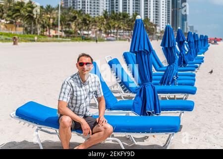 Junge Mann Mode Millennial in roten Sonnenbrillen am Strand an sonnigen Tag in Miami, Florida sitzen auf blauen Liegestühlen, Sonnenschirme von Hotel oder Wohnung c Stockfoto