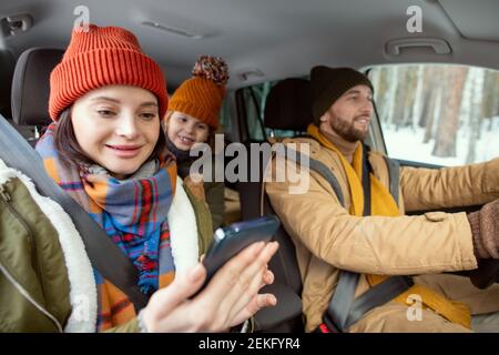 Glückliche junge Mutter in warmer Winterkleidung Blick auf den Bildschirm Smartphone gegen ihre kleine Tochter, die auf dem Rücksitz des Autos sitzt Während der Fahrt Stockfoto