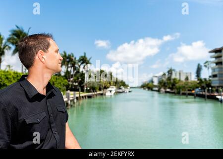 Porträt eines jungen Mannes in schwarzem Hemd auf einer Brücke in Bal Harbour, Miami Florida mit grünem Ozean Biscayne Bay und Wohngebäuden Stockfoto