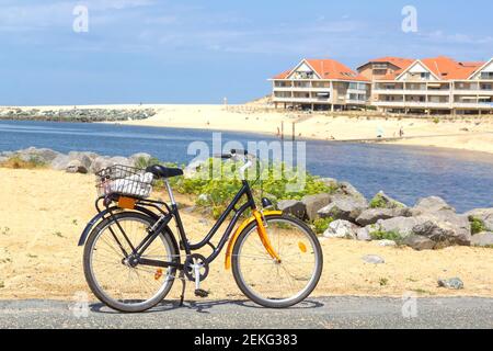 Orange Fahrrad mit der schönen Landschaft des Landes im Vordergrund: Blaues Meer, Sandstrand und Ferienhäuser mit typischen orange Ziegeldächern Stockfoto