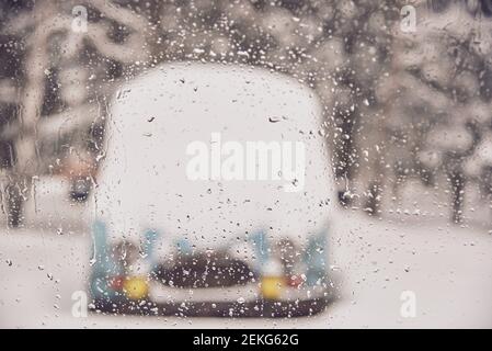 Durch das nasse Glas sieht man die Silhouette eines schneebedeckten Minivans. Geschmolzener Schnee auf dem Fenster. Stockfoto