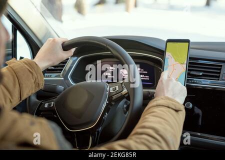 Hände des jungen Mannes in warmer Winterkleidung halten von Steer Während Sie im Auto sitzen und auf die Karte auf dem Bildschirm zeigen Smartphone während der Reise Stockfoto