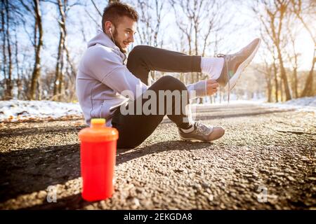 Seitenansicht der jungen zufrieden motiviert und konzentriert sportlich aktiv Mann in Wintersportbekleidung zieht Sneaker an, während er innen sitzt Der verschneite Wald auf dem Ro Stockfoto