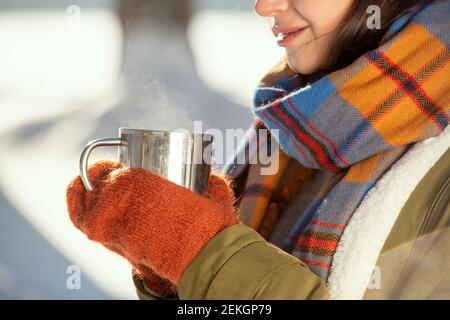 Hände der jungen Frau in warmer Jacke, Schal und Wollgestrickte Fäustlinge halten metallischen Becher mit heißem Tee, während vor der Kamera stehen Stockfoto