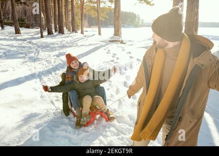 Glücklicher junger Vater in warmer Winterkleidung zieht Schlitten mit seinem Lachende Frau und ihre süße kleine Tochter, während Spaß Am Wintertag Stockfoto