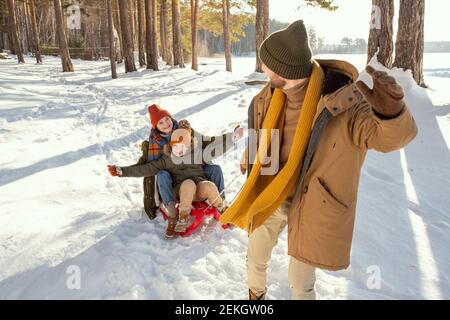 Fröhlicher junger Mann in warmer Winterkleidung zieht Schlitten mit seinem Lachende Frau und ihre süße kleine Tochter, während Spaß Am Wintertag Stockfoto