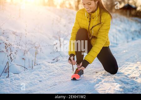 Nahaufnahme Ansicht der glücklich attraktive fleißiges aktives Fitness-Mädchen Mit Kopfhörern im Winter Sportkleidung binden Schnürsenkel auf Turnschuhe draußen Im Schnee natu Stockfoto