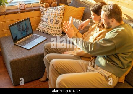 Glücklich junge liebevolle Paar in Casualwear winken Hände zum Mann Und seine niedliche kleine Tochter auf Laptop-Display während online Kommunikation Stockfoto