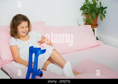 Ein fröhliches Teenager-Mädchen liegt auf dem Bett, nachdem ihr Bein in der Notaufnahme gegossen wurde. Das Kind hat ein gebrochenes Bein, einen gebrochenen Knochen Stockfoto