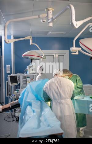 Der Patient ist mit einem Tuch bedeckt, das auf dem Operationstisch liegt, während sich zwei professionelle Chirurgen über ihn beugen, weibliche Assistentin, die Daten steuert Stockfoto