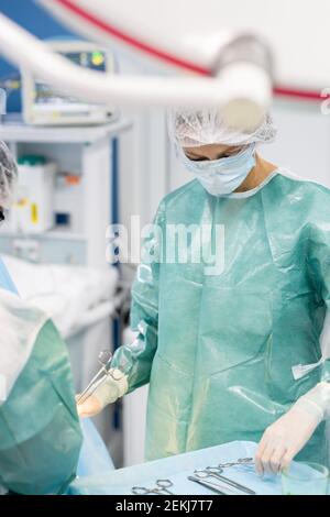 Junge Assistentin des professionellen Chirurgen in schützende Arbeitskleidung stehend Durch den Patienten während der Operation und Einnahme von medizinischem Instrument Stockfoto