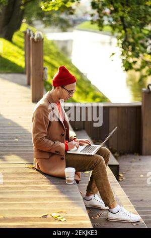 Lächelnder Freiberufler Mann tragen Brille reden auf Handy, arbeiten auf Laptop, sitzen im Freien. Stockfoto