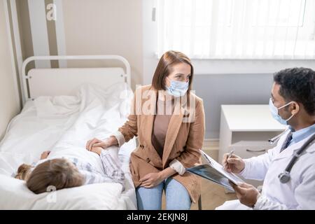 Junge ernsthafte Brünette weiblich in Schutzmaske im Gespräch mit Arzt Über ihre kranke Freundin, die im Bett in der Nähe liegt Modernes Krankenhaus Stockfoto