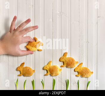 Eine Kinderhand spielt mit Cookies in Form von Kaninchen, die auf dem grünen Gras galoppieren. Blick von oben auf weiß mit vertikalen Linien Holzhintergrundzaun. Stockfoto