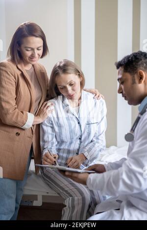 Zuversichtlich männlichen Arzt in Whitecoat hält medizinische Dokument, während seine Patientin, die ihre Unterschrift setzt und ihre Freundin in der Nähe steht Von Stockfoto