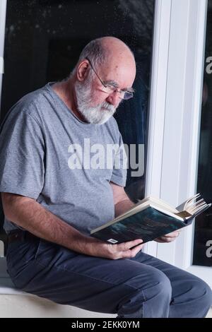 Älterer Mann, der nachts in seinem Wintergarten sitzt und einen Roman liest. Stockfoto
