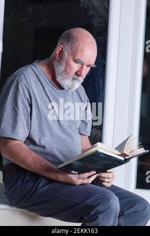 Älterer Mann, der nachts in seinem Wintergarten sitzt und einen Roman liest. Stockfoto
