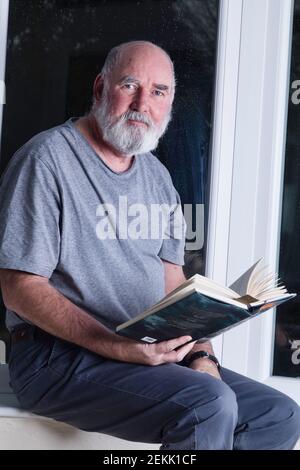 Älterer Mann, der nachts in seinem Wintergarten sitzt und einen Roman liest. Stockfoto