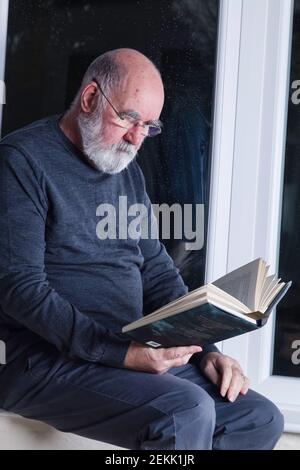 Älterer Mann, der nachts in seinem Wintergarten sitzt und einen Roman liest. Stockfoto