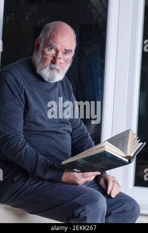 Älterer Mann, der nachts in seinem Wintergarten sitzt und einen Roman liest. Stockfoto