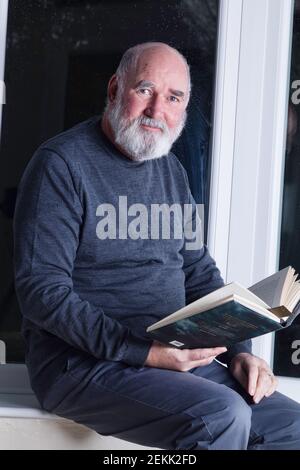 Älterer Mann, der nachts in seinem Wintergarten sitzt und einen Roman liest. Stockfoto
