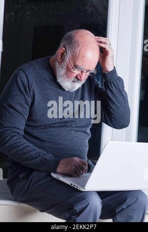 Ein älterer Mann, der nachts in seinem Wintergarten sitzt und versucht, einen Laptop zu benutzen. Stockfoto
